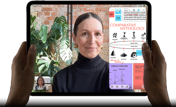 iPad Pro i space black vist forfra i vandret position, afrundede hjørner, sort skærmkant, et par hænder holder enheden på venstre og højre side, skærmen viser et FaceTime-opkald, der oversættes live, en uddannelsesapp viser en præsentation af “Comparative Mythology” med tekst, diagrammer og håndskrevne noter